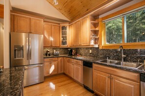 Cocina con armarios de madera, electrodomésticos de acero inoxidable, encimeras de granito y ventana con vistas a árboles.