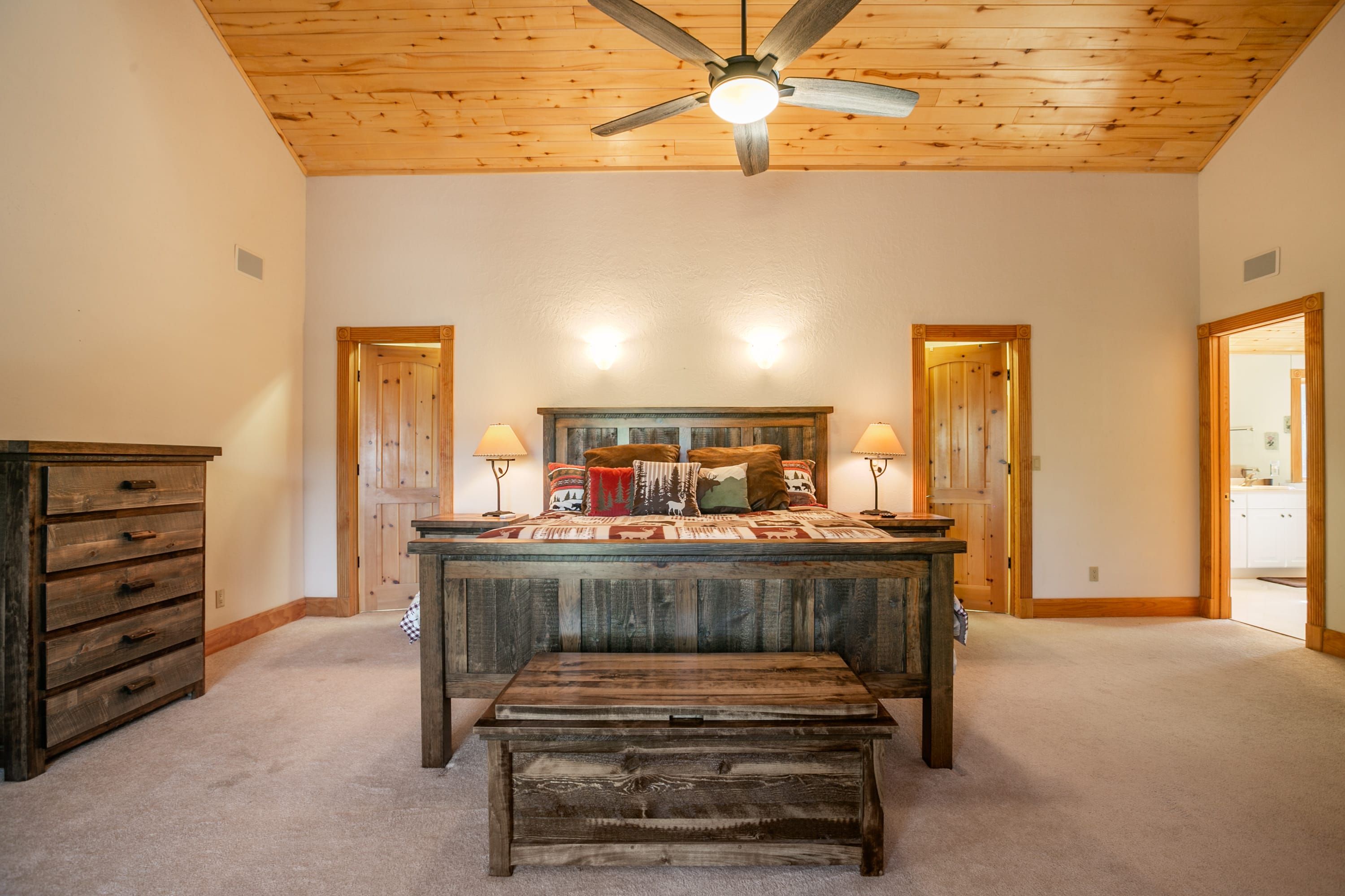 A spacious room with a wooden ceiling fan, a large bed with a wooden headboard, two bedside lamps, and a wooden bench.