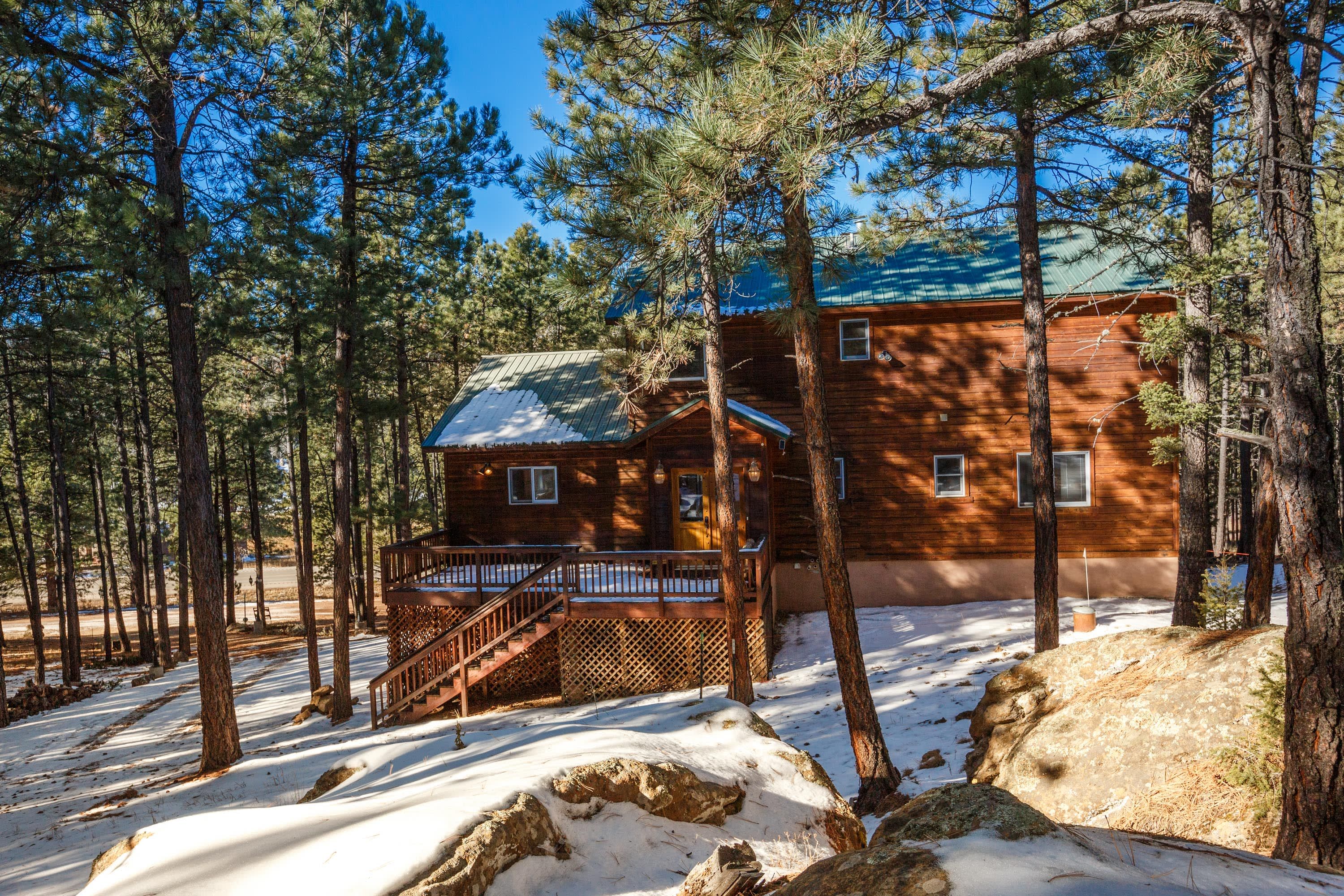 A log cabin with a deck and stairs, surrounded by tall pine trees and snow-covered ground.
