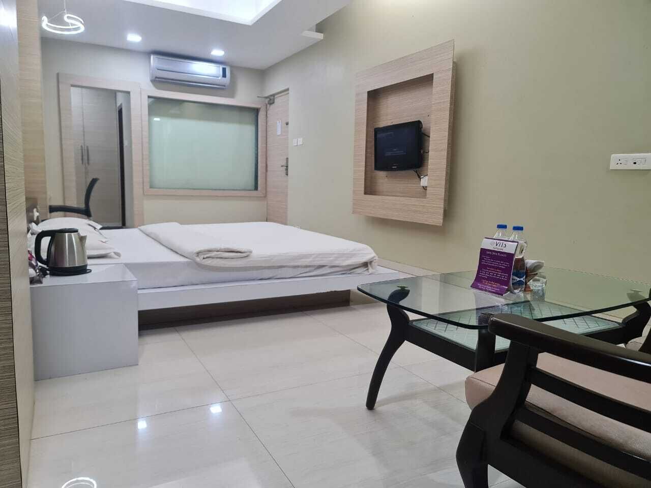 A hotel room with a bed, a glass table, a coffee maker, a TV, and a small wooden shelf.