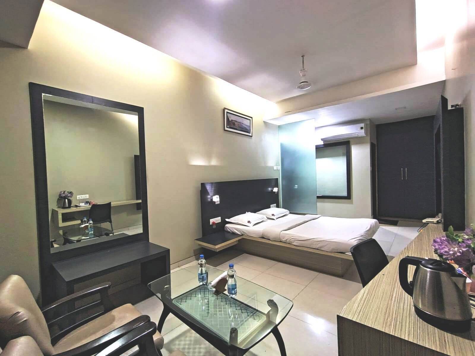 A hotel room with a bed, a desk with a chair, a large mirror, a small table with two bottles, and a kettle on a wooden table.