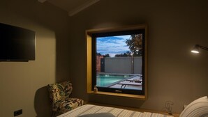 Un dormitorio con cama, ventana con vista a una piscina exterior, una silla y televisor en la pared.