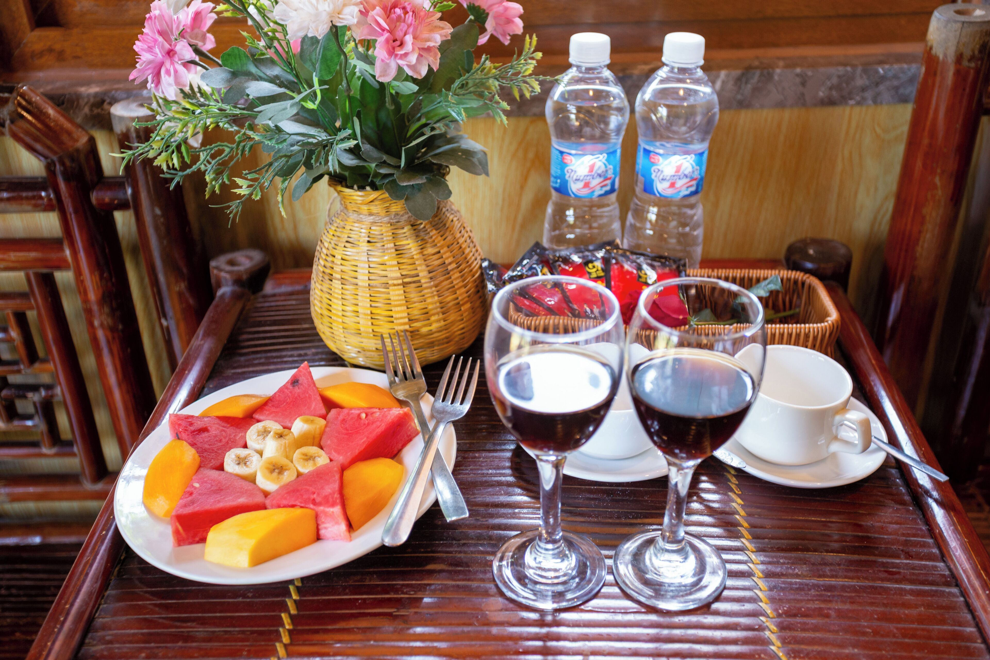 A wooden table with a plate of fruit, two wine glasses, a vase with flowers, and two water bottles.