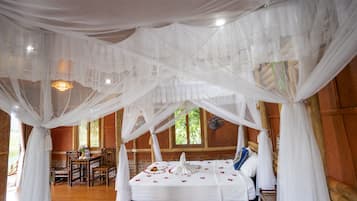A room with a bed under a canopy, a dining table with chairs, and a view of greenery outside.