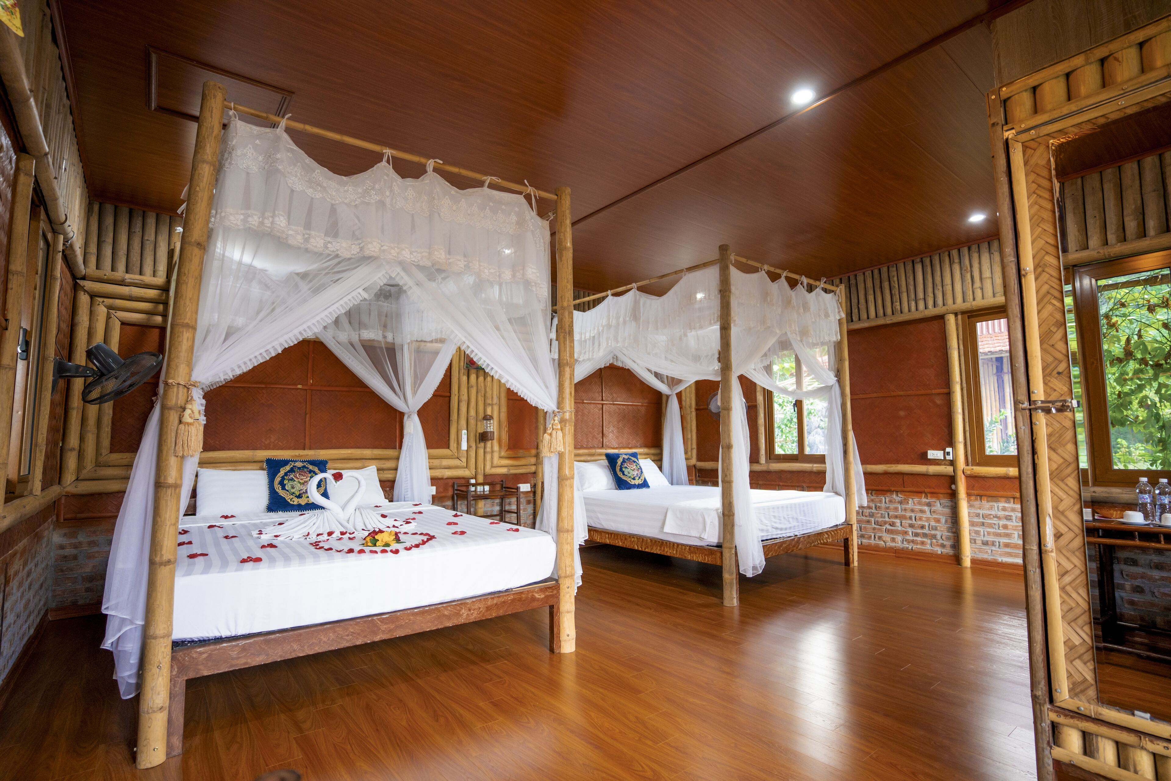 A room with two wooden four-poster beds with white canopies, wooden floors, and a brick wall.