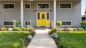 A two-story building with a yellow front door, a covered walkway, and a garden with various plants. - Ultra Comfort + Style! HOT TUB & Fireplace! Two Living Rooms >10 min to Downtown & BSU! (Boise)
