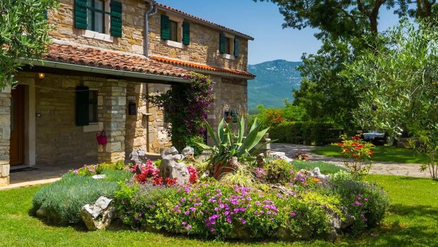 A stone building with a tiled roof, a garden with colorful flowers and plants, and a mountainous backdrop.