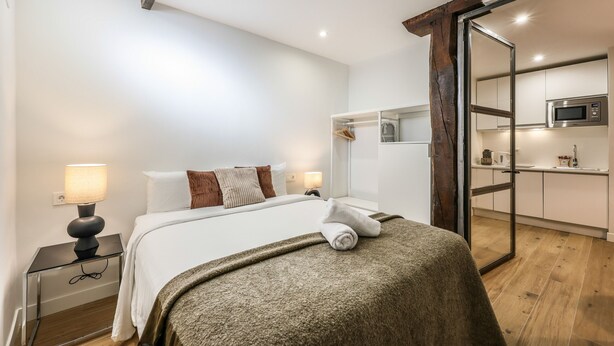 A modern hotel room with a bed, bedside lamps, a wooden floor, a glass door leading to a kitchenette, and a white wall.
