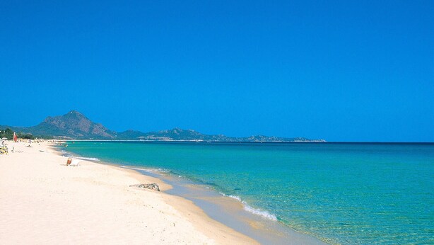 Ein Strand mit klarem türkisblauem Wasser, einem sandigen Ufer und einem Berg im Hintergrund.