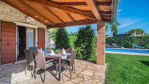 An outdoor patio with wicker furniture and a stone floor, a covered area with a wooden ceiling, and a view of a swimming pool and greenery in the background. - Beautiful home in Marcana (Marcana)