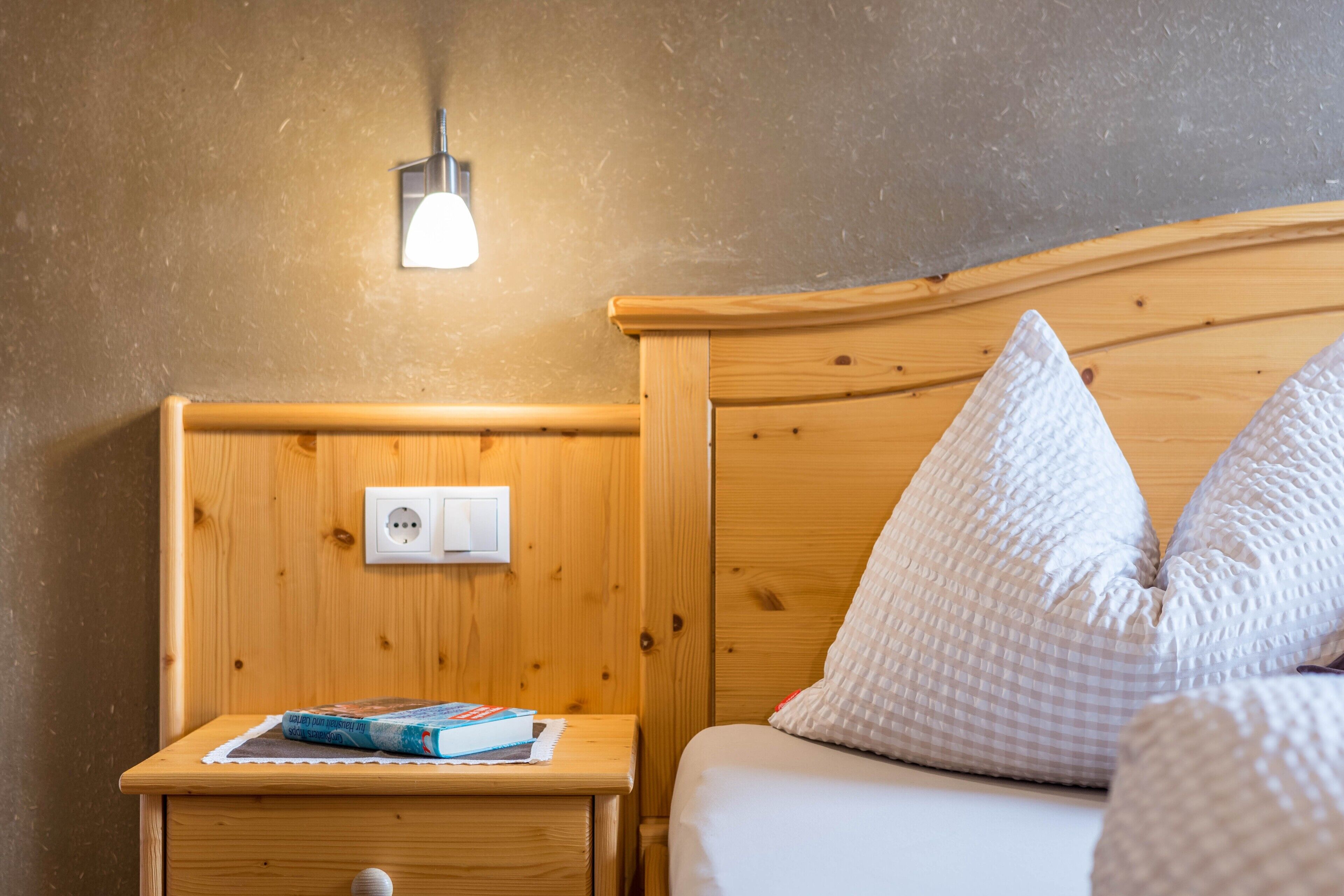 A wooden bed with white pillows, a bedside table with a book, a wall socket, and a wall-mounted light fixture.