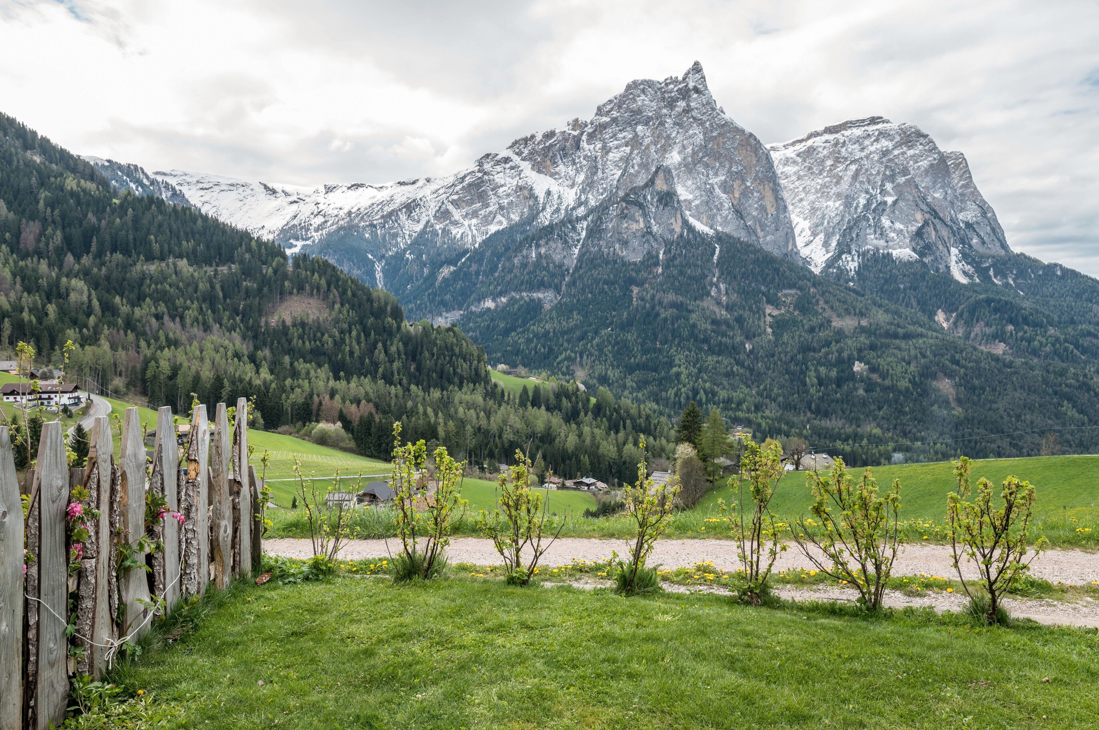 A mountainous landscape with a wooden fence, green fields, and a house in the foreground.
