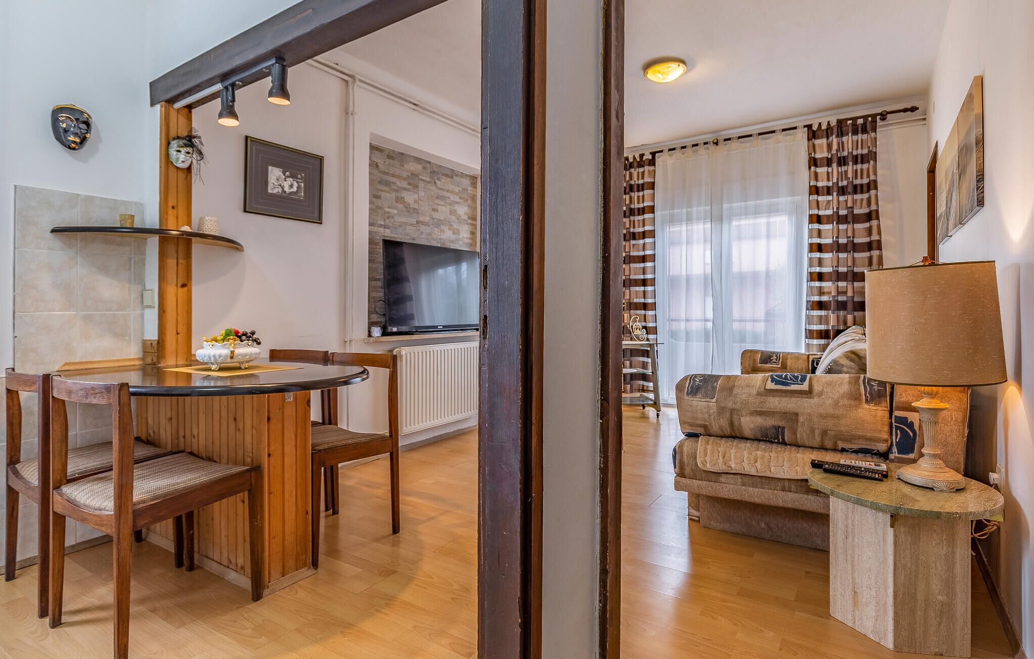 Een eetgedeelte met houten stoelen en een tafel, een woonkamer met een bank en een lamp, en een keuken met een televisie en een betegelde achterwand.