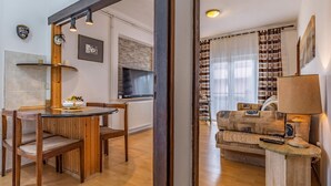 Een eetgedeelte met houten stoelen en een tafel, een woonkamer met een bank en een lamp, en een keuken met een televisie en een betegelde achterwand.