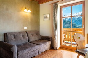 Una habitación acogedora con suelo de madera, un sofá gris, una silla de madera, una ventana con vistas a montañas y una lámpara fijada a la pared.