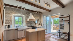 Una cocina con paredes de piedra, vigas de madera y una isla grande. Hay una ventana con vista a árboles, un lavamanos y una estufa.