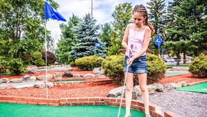 A miniature golf course with a green playing surface, a blue ball, and a player in a pink top and denim shorts.