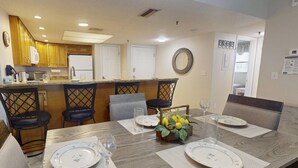 A dining area with a wooden table set for four, surrounded by chairs. A kitchen with wooden cabinets and a refrigerator is visible in the background.