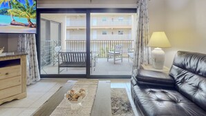 A living room with a black leather sofa, a TV on the wall, a balcony with a view of other buildings, and a lamp on a side table.