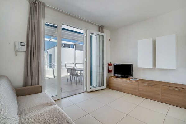 A modern room with a sofa, a TV on a wooden cabinet, a sliding glass door to a balcony, and a fire extinguisher on the wall.