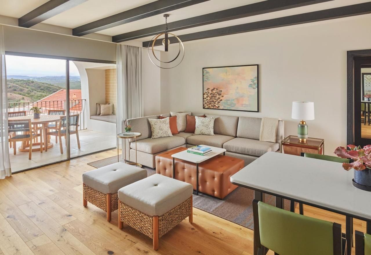 Uma sala de estar espaçosa com um sofá grande, pufes, uma mesa de centro e um espaço para refeições. Há uma varanda com vista para o campo.