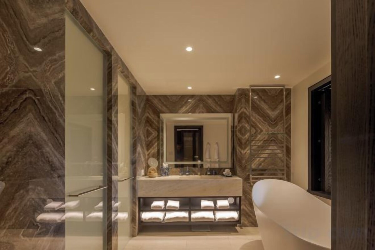 A modern bathroom with a marble wall, a built-in bench with towels, a large mirror, and a freestanding bathtub.