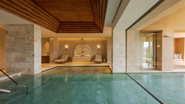 Une piscine intérieure moderne avec un plafond en bois, des murs en marbre et un espace bien-être équipé de deux chaises longues.