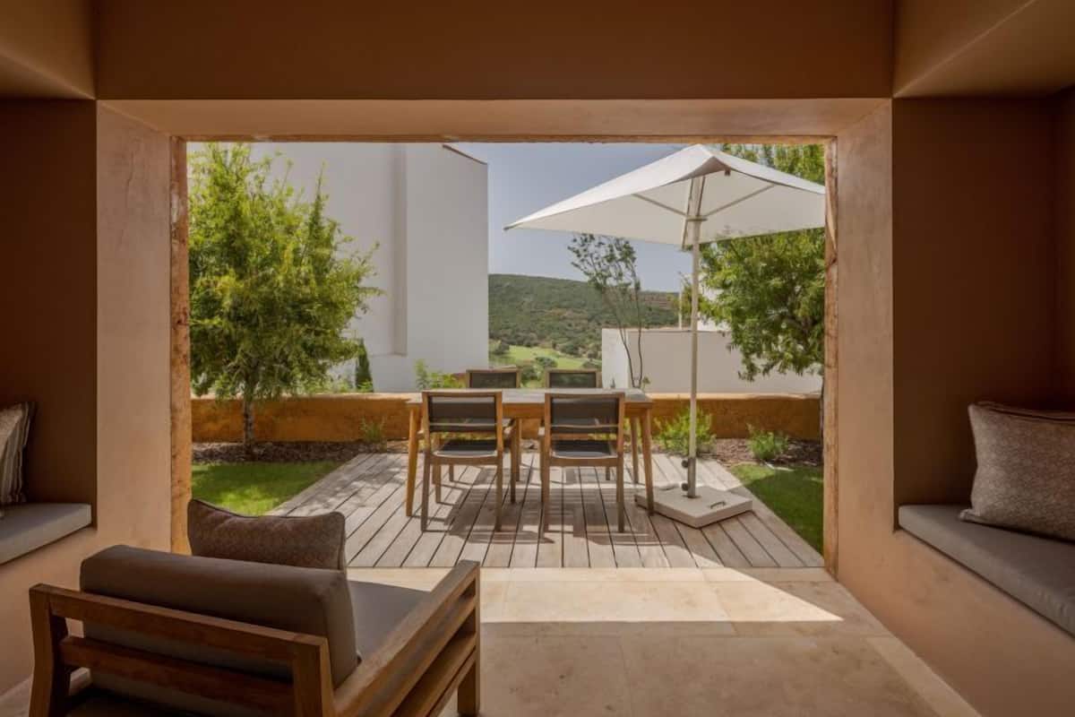 A modern patio with a wooden table and chairs, a cushioned bench, and a white umbrella.