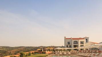 A pool with lounge chairs and umbrellas, a large building with a red roof, and a hilly landscape in the background.