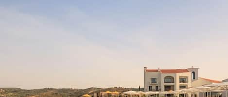 Una piscina con lettini e ombrelloni, un edificio con tetto rosso e un paesaggio collinare sullo sfondo.