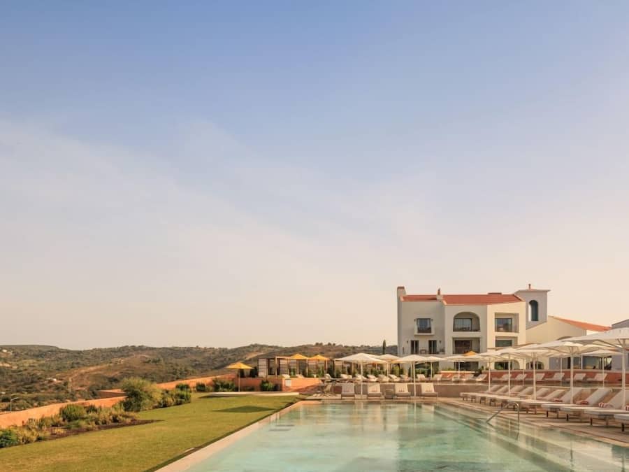 A pool with lounge chairs and umbrellas, a large building with a red roof, and a hilly landscape in the background.