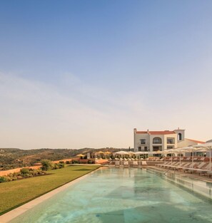 Una piscina con sillones de descanso y sombrillas, un edificio grande con techo rojo y un paisaje montañoso al fondo.