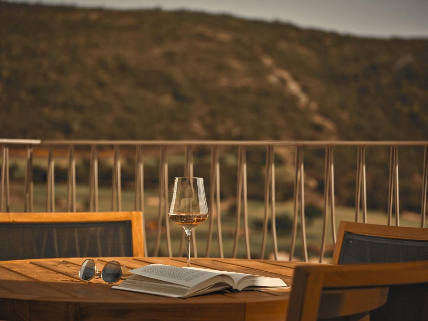 A wooden table with a book, a pair of sunglasses, and a glass of wine on a balcony with a view of a mountainous landscape.