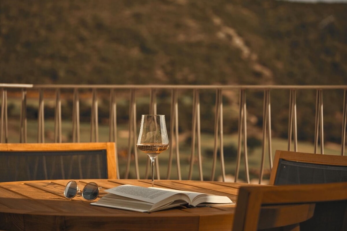 Mesa de madeira com um livro, um par de óculos de sol e um copo de vinho numa varanda com vista para uma paisagem montanhosa.