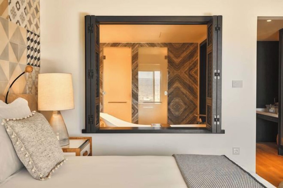 A hotel room with a bed, a nightstand, a lamp, and a large mirror reflecting the room's interior.