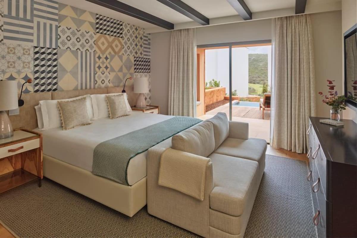 A modern hotel room with a large bed, a sofa, a desk, and a view of the outdoors.