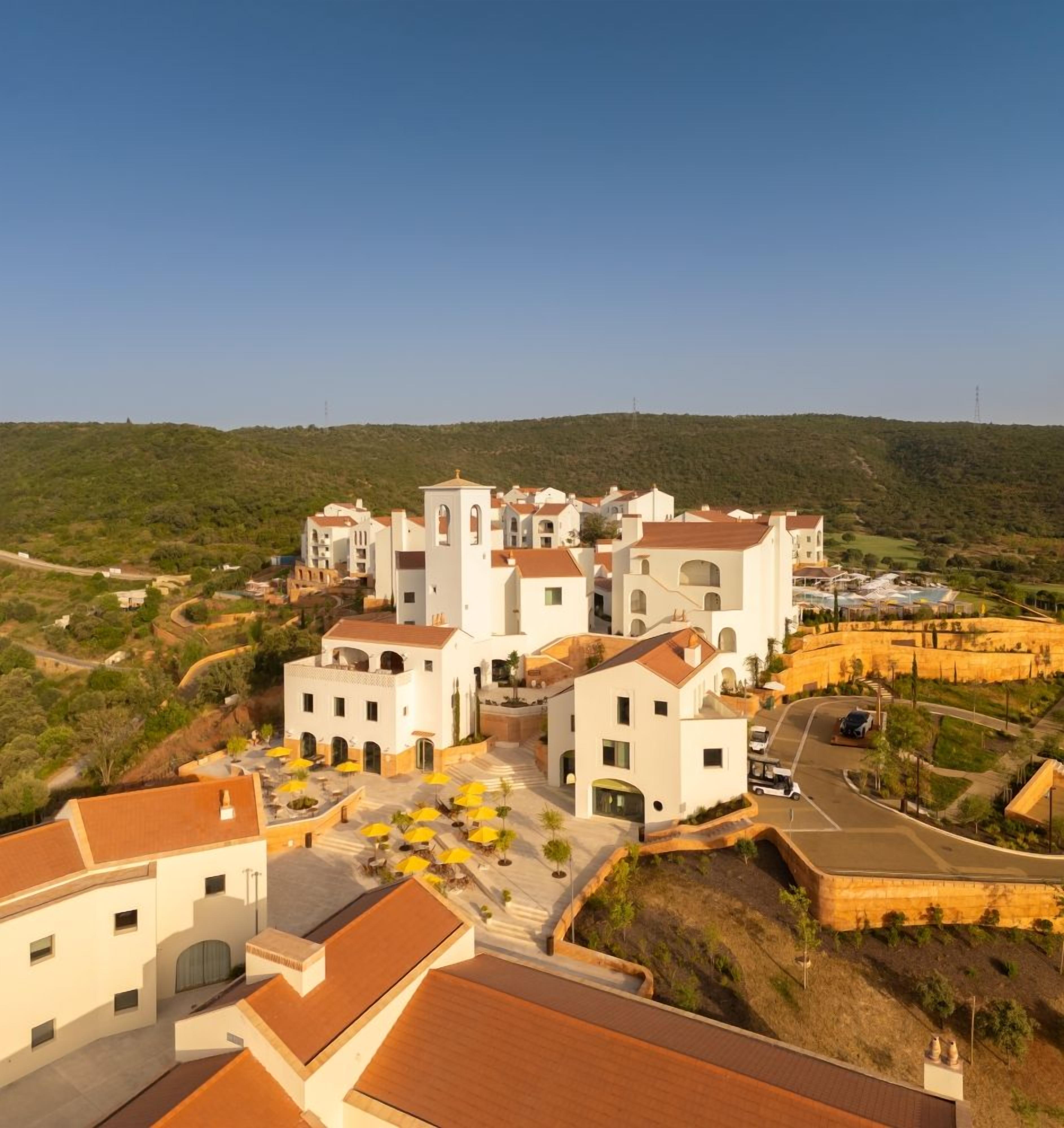 A coastal village with white buildings, a central tower, and a paved area with yellow umbrellas.
