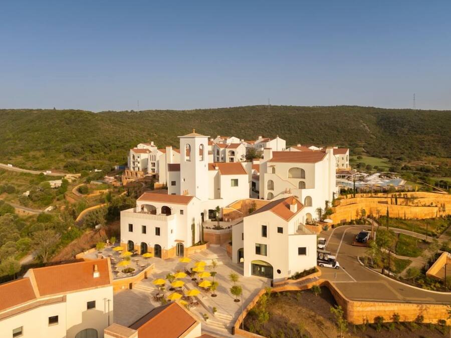 A coastal village with white buildings, a central tower, and a paved area with yellow umbrellas.