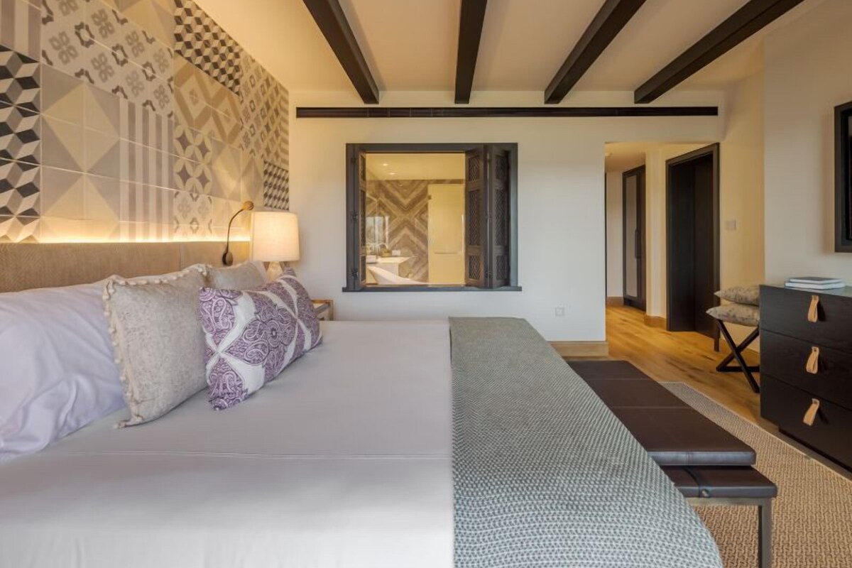 Une chambre moderne avec un grand lit, un banc, une commode et un mur orné d’un motif.