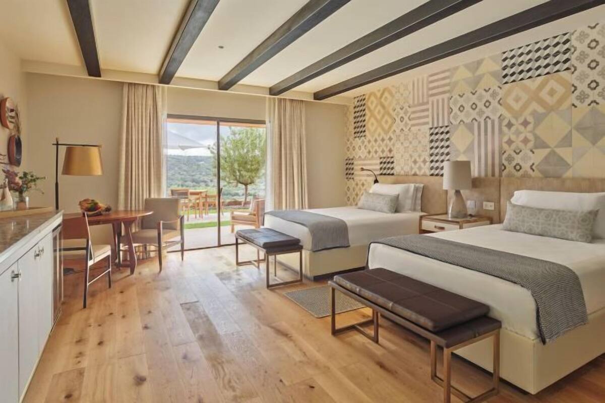 A hotel room with a bed, a desk, a chair, a bench, and a view of the outdoors.