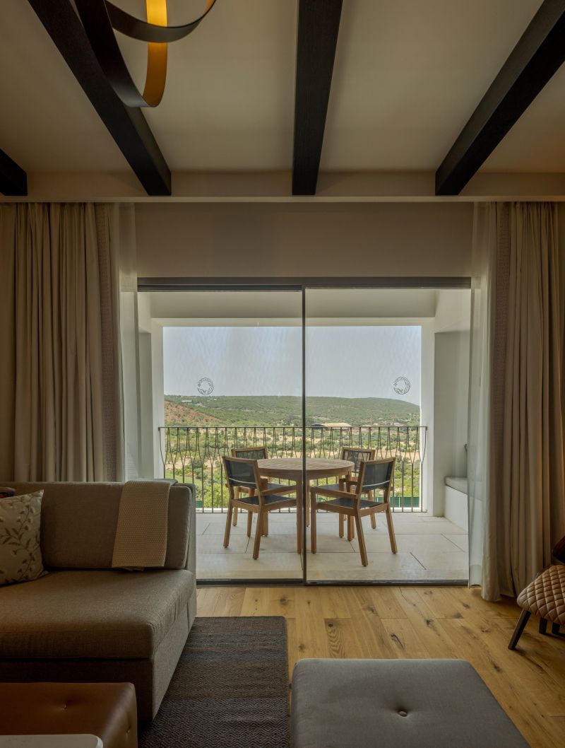 Een moderne woonkamer met een bank, een salontafel en een eethoek met een tafel en stoelen. Een grote schuifpui leidt naar een terras buiten met uitzicht op groene heuvels.