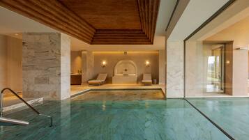 A modern indoor swimming pool with a wooden ceiling, marble walls, and a spa area with two sun loungers.