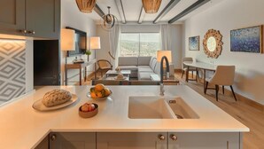 Une cuisine moderne dotée d’un îlot central, un coin repas avec une table et des chaises, et un salon avec un canapé et une vue sur l’extérieur.