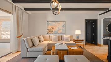 A modern living room with a sectional sofa, ottomans, a coffee table, and a pendant light. There is a painting on the wall and a door leading to another room.