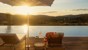 Une terrasse en bois avec deux chaises longues, une table sur laquelle se trouve un verre et un panier de citrons, donnant sur une étendue d’eau avec un coucher de soleil en arrière-plan.