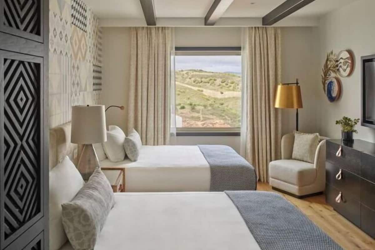 A hotel room with two beds, a desk, a chair, a lamp, and a large window with a view of a landscape.