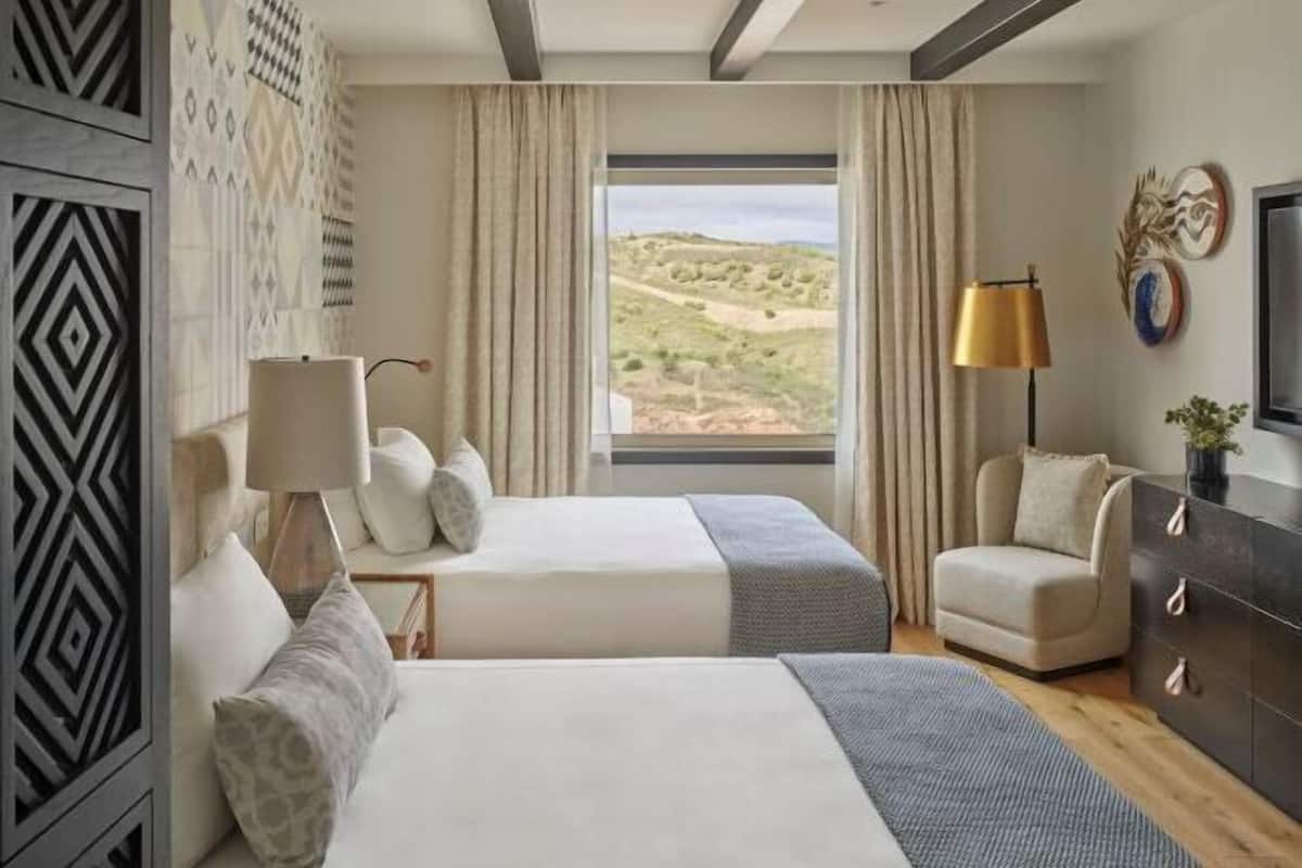 A hotel room with two beds, a desk, a chair, a lamp, a dresser, and a large window with a view of a landscape.