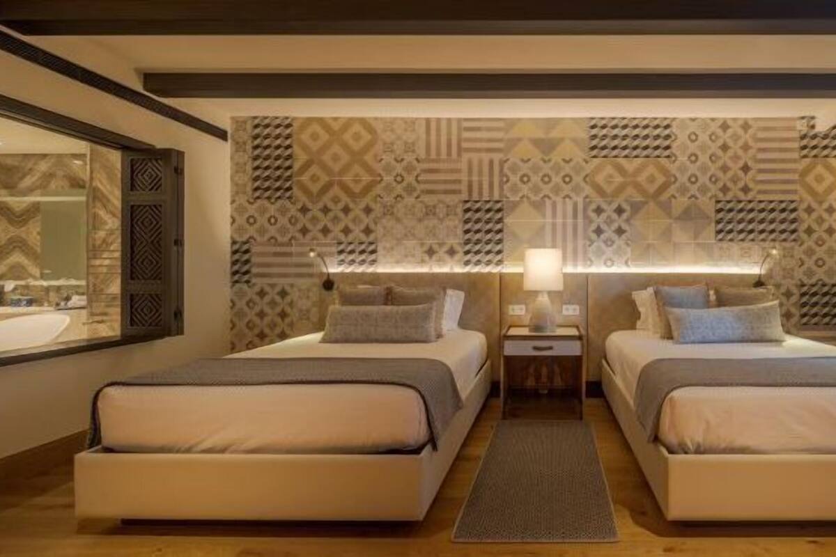 A hotel room with two beds, a large mirror, patterned wallpaper, and a nightstand with a lamp.