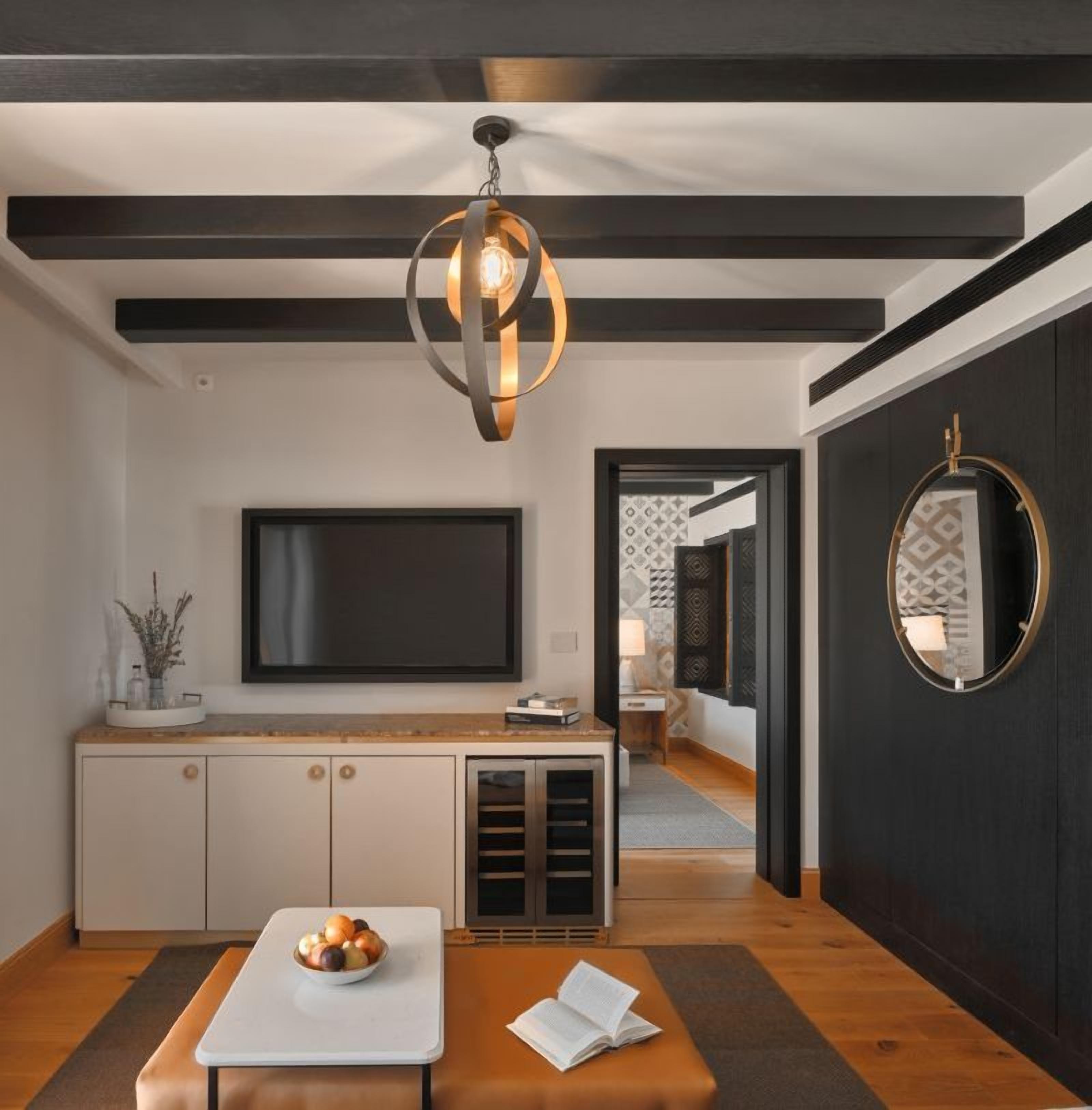 A modern living room with a flat-screen TV, a wooden bench, a bowl of fruit, and a unique pendant light.