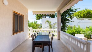 Un espace extérieur abrité, aménagé pour les repas, avec une table et des chaises, une rambarde blanche et de la verdure en arrière-plan.